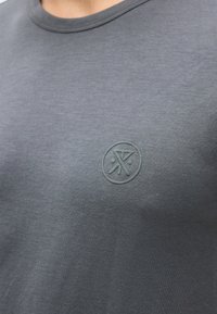 Σκούρο γκρι μπλουζάκι με στρογγυλή λαιμόκοψη και διακριτικό ανάγλυφο κυκλικό λογότυπο στο αριστερό στήθος με αφηρημένα γεωμετρικά σχήματα.