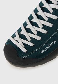 Zielony zamszowy but wspinaczkowy z białymi sznurówkami, czarnym gumowym czubkiem i detalami szwalniczymi po bokach; nazwa marki "SCARPA" nadrukowana białym tekstem.