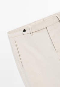 Beige broek van zacht materiaal, met een platte voorkant, zwarte knoopsluiting en schuine zijzakken. Glad oppervlak zonder patronen.