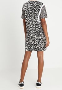 Osoba oblečená v šatech s krátkým rukávem v černobílem vzoru s bílými detaily, v černobílých teniskách, stojící před bílým pozadím.