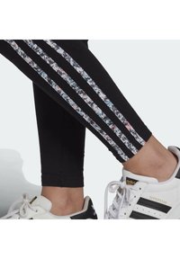 Svarta leggings har tre vertikala ränder med ett färgglatt blommönster. De är kombinerade med vita och svarta Adidas-sneakers.