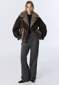 Femme se tenant debout portant une veste en shearling brun foncé, un haut noir, un pantalon large gris et des chaussures à talons pointues noires sur un fond blanc.