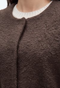 Gros plan d'une personne portant une veste marron texturée par-dessus un pull blanc à côtes avec col rond.