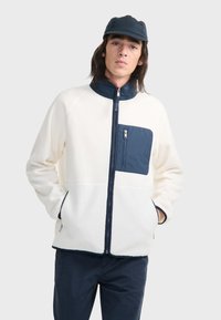 Fleecejacke in Creme mit marineblauen Akzenten. Verfügt über einen hohen Kragen, Reißverschlusstaschen und eine kontrastierende Brusttasche. Weicher Stoff und lockere Passform.