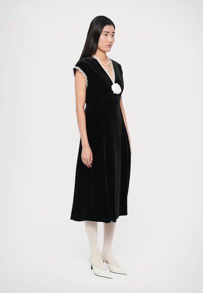 Vestido de veludo preto com acabamento em renda, decote em V e detalhe de flor branca no peito. Usado com collants creme e sapatos brancos pontiagudos.