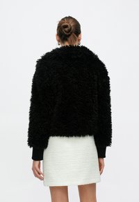 Μαύρο κοντό σακάκι από ψεύτικη γο ύνα με μακριά μανίκια και υφή, συνδυασμένο με λευκή υφασμάτινη φούστα. Άποψη από πίσω.