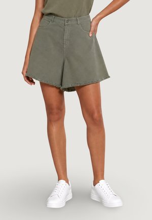 Donna che indossa pantaloncini larghi verde oliva con orli sfilacciati, una maglia oliva e sneakers bianche, in piedi con una mano in tasca.