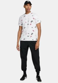 T-shirt bianca con loghi Nike sparsi in rosso, nero e grigio, abbinata a pantaloni della tuta neri e scarpe sportive nere.