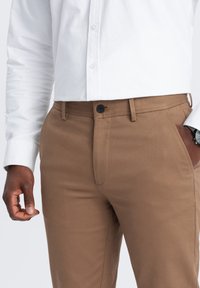 Pantaloni marroni realizzati in tessuto testurizzato con chiusura a bottone e tasche laterali, abbinati a una camicia bianca con colletto.