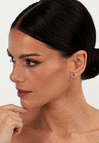 Goldene Ohrstecker mit kleinen Diamantakzenten, vor einem neutralen Hautton präsentiert, wodurch der Glanz der Ohrringe und die zarten Züge des Modells hervorgehoben werden.