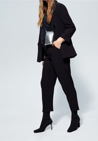 Blazer noir sur mesure à coupe décontractée, associé à un pantalon noir ajusté et des bottines style chaussette. Une pochette métallique argentée apporte un contraste.