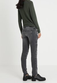 Mujer vestida con una blusa de manga larga verde oscura, vaqueros ajustados gris con detalles de cremallera lateral y zapatos de cuero negros de suela gruesa, vista desde atrás.