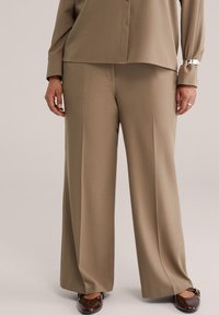 Bež široke hlače iz gladkega blaga z izrazitim krojem, s ploskim sprednjim delom in stranskimi žepi, združene z bleščečimi rjavimi čevlji.