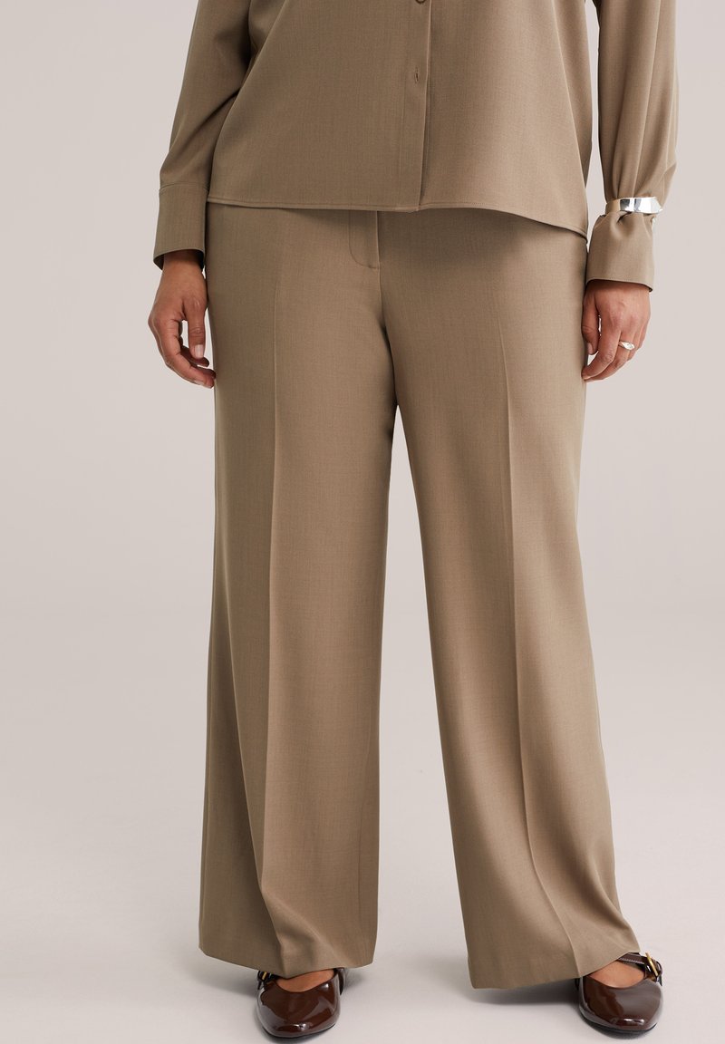 Bež široke hlače iz gladkega blaga z izrazitim krojem, s ploskim sprednjim delom in stranskimi žepi, združene z bleščečimi rjavimi čevlji.