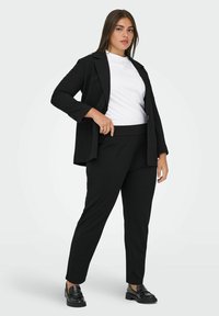 Black suit jacket with a lapel, paired with black trousers. Model wears a white t-shirt. Black loafers complete the outfit.