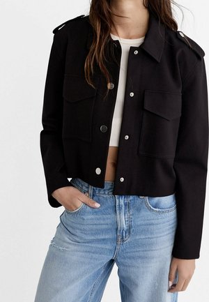 Vrouw draagt een korte zwarte jas met zilveren knopen over een witte cropped top en lichtblauwe hooggesloten jeans, hand in de zak.