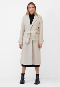 Beige belted coat with a textured finish, notched lapels, and pockets, worn over a black dress and paired with black ankle boots.