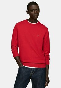 Maglione rosso a maglia con collo rotondo e polsini a coste, con un piccolo logo sul petto. Abbinato a jeans in denim blu scuro.