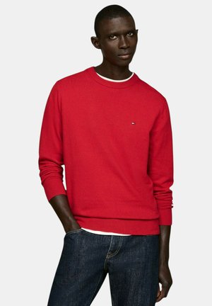 Roter Strickpullover mit Rundhalsausschnitt und gerippten Bündchen, ausgestattet mit einem kleinen Logo-Detail auf der Brust. Kombiniert mit dunkelblauen Jeans.