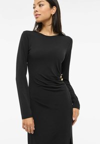 Juoda ilgas rankovėmis suknelė iš tamprios medžiagos, pasižyminti drapiruotu dizainu su auksiniais mygtukų akcentais juosmens srityje ir šoniniu plyšiu.