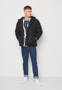 Svart boblejakke med hette, glidelås foran, horisontal søm, kombinert med en blå og hvit mønstrete skjorte, denimjeans og hvite sneakers.