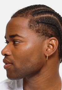 Goldene Creolen an einer Person mit geflochtenem Haar, die eine strukturierte Hautfarbe und glatte Gesichtszüge mit natürlichen Highlights zeigt.