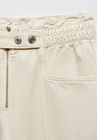 Pantaloni bej deschis cu talie elastică, nasturi metalici argintii și fermoar. Materialul are o textură netedă și un buzunar lateral.