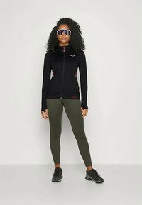 Vrouw staand, gekleed in een zwarte zip-up jas, olijfgroene leggings, zwarte sneakers en grote reflecterende sportzonnebril, tegen een effen achtergrond.