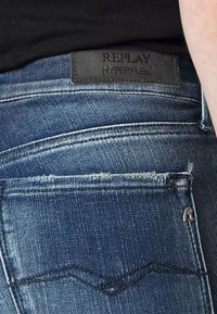 Vaqueros de mezclilla azul con una parche de cuero negro "REPLAY HYPERFLEX". Presentan un lavado desvanecido, un ligero efecto de arrugado, y un diseño clásico de bolsillo trasero.