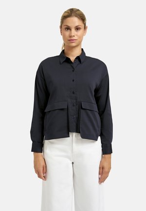 Woman with blonde hair pulled back, wearing a black button-up shirt with flap pockets and white pants, standing against a plain background.