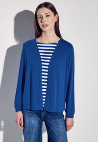 Blå lättviktskofta med öppen front, långa ärmar, ihop med en vit och blå randig skjorta under. Denimjeans syns.