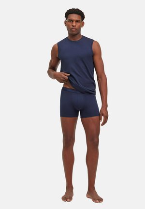 Homme portant un débardeur bleu marine et un boxer, soulevant légèrement son t-shirt pour montrer la ceinture, debout pieds nus sur fond blanc.