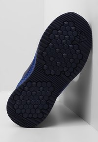 Sole obuvi s texturovanou tmavě modrou gumou a hexagonálním vzorem, s označením bez zanechání stop, nakloněná proti bílému pozadí.