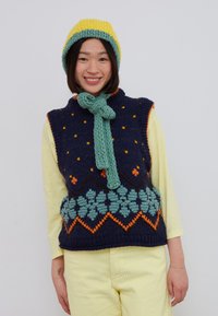 Personne souriante portant un bonnet jaune, une écharpe verte, un gilet en tricot bleu marine avec motif floral, une chemise à manches longues jaune et un pantalon jaune devant un mur blanc.