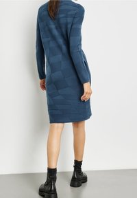 Robe texturée bleue avec manches longues et silhouette ajustée, associée à des bottines noires. Accentuée d'un motif.