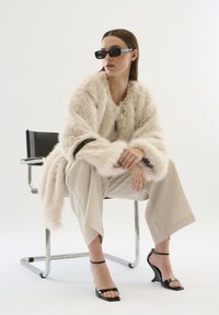 Faux-Fur-Mantel in Beige, weit geschnittene cremefarbene Hose, schwarze Riemchen-Heels und rechteckige Sonnenbrille. Model sitzt auf einem Chromstuhl.
