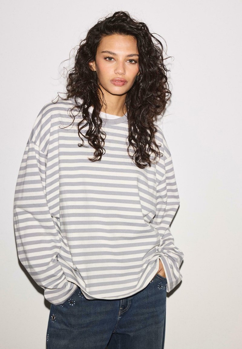 Nainen, jolla on pitkät, kiharat tummat hiukset, päällään väljä harmaa-valkoraidallinen pitkähiha paita ja tummat farkut, kädet taskuissa yksiväristä taustaa vasten.