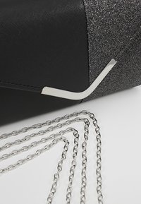 Černé psaníčko s třpytivými stříbrnými akcenty, texturovaným povrchem a stříbrným řetízkovým popruhem. Má elegantní, úhlový design.