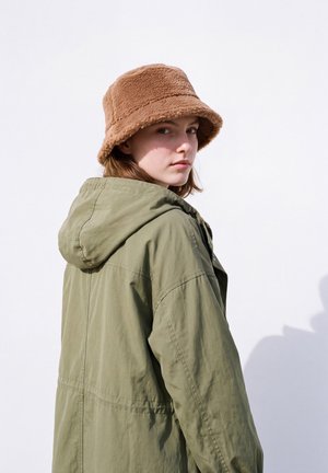 Une femme portant un bob marron en polaire et une veste à capuche vert olive regarde par-dessus son épaule sur un fond blanc uni.