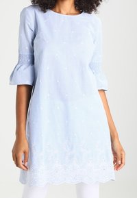 Tunique rayée bleu clair avec manches bouffantes, ornée de broderies florales et d'un détail d'ourlet festonné. Fabriquée en tissu respirant.