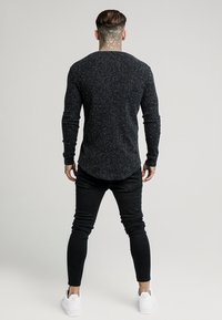 Černá texturovaná košile s dlouhým rukávem a zakřiveným lemem, v kombinaci s černými úzkými kalhotami a bílými teniskami. Pohled zezadu.