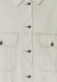 Ljusgrå denimskjorta med spetsig krage, snedstygn och svarta metallknappar. Har två framfickor med lock.