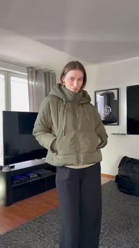 Casaco acolchoado verde-oliva com capuz ajustado, fecho de correr e cordão, mangas largas e dois bolsos frontais, usado por cima de calças pretas.