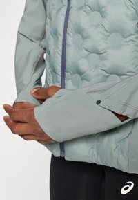 Svetlozelená izolačná bunda s okrúhlym prešívaným vzorom; má zips v prednej časti, kontrastný fialový zips a manžety s otvorom na palec pre lepšiu ochranu.