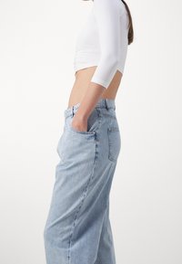 Jeans de mezclilla azul claro con un ajuste relajado, que presentan un diseño clásico de cinco bolsillos y costuras visibles. Combinados con una blusa blanca corta.