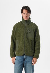 Chaqueta de forro polar verde con un acabado texturizado, cuello alto, cierre frontal con cremallera y múltiples bolsillos con cremallera. Acentos de tela en contraste.