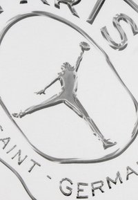 Λευκό ύφασμα με ανάγλυφο, λαμπερό ασημί λογότυπο Jordan και κείμενο «PARIS SAINT-GERMAIN», που απεικονίζει έναν παίκτη μπάσκετ στον αέρα.