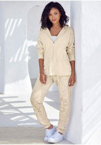 Beige Zip-Hoodie mit Kordelzug, kombiniert mit passenden Hosen mit dezenten Blumenmustern. Weiße Sneaker runden das Outfit ab.