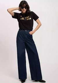 Czarna koszulka z złotym napisem, wysokie szerokie dżinsy w niebieskim kolorze oraz czarne okulary przeciwsłoneczne. Gładka faktura, casualowy design, nowoczesne dopasowanie.