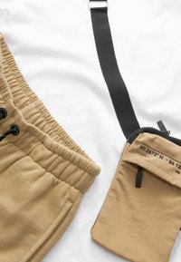 Pantalones de chándal caqui con cinturilla elástica, acompañados de un estuche con cremallera color beige y una correa negra con coordenadas impresas. Fondo blanco.